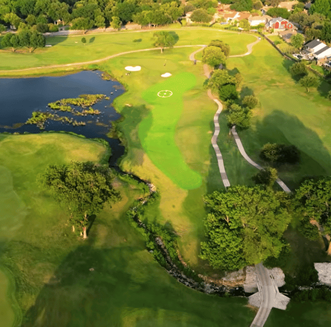 Aerial view of a green bordered by a pond and bunkers, with a creek and tree-lined fairway below.